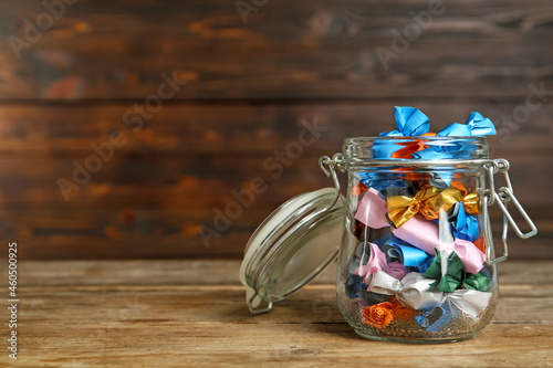 Glass jar with candies in colorful wrappers on wooden table. Space for text