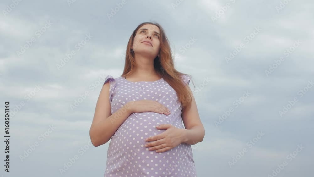 dreaming pregnant woman looks up to sky with gratitude in field on background of beautiful cloudy sky