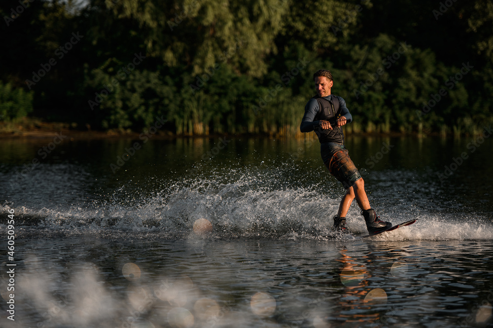 Active sportive man in wetsuit energetically balancing on water surface on wakeboard Active sportive man in wetsuit energetically balancing on water surface on wakeboard