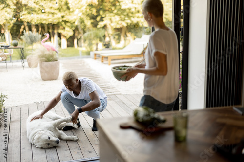 Black man caress dog while his european girlfriend looking on it and holding bowl with salad. Concept of relationship. Modern domestic lifestyle. Idea of healthy eating. Couple at home terrace