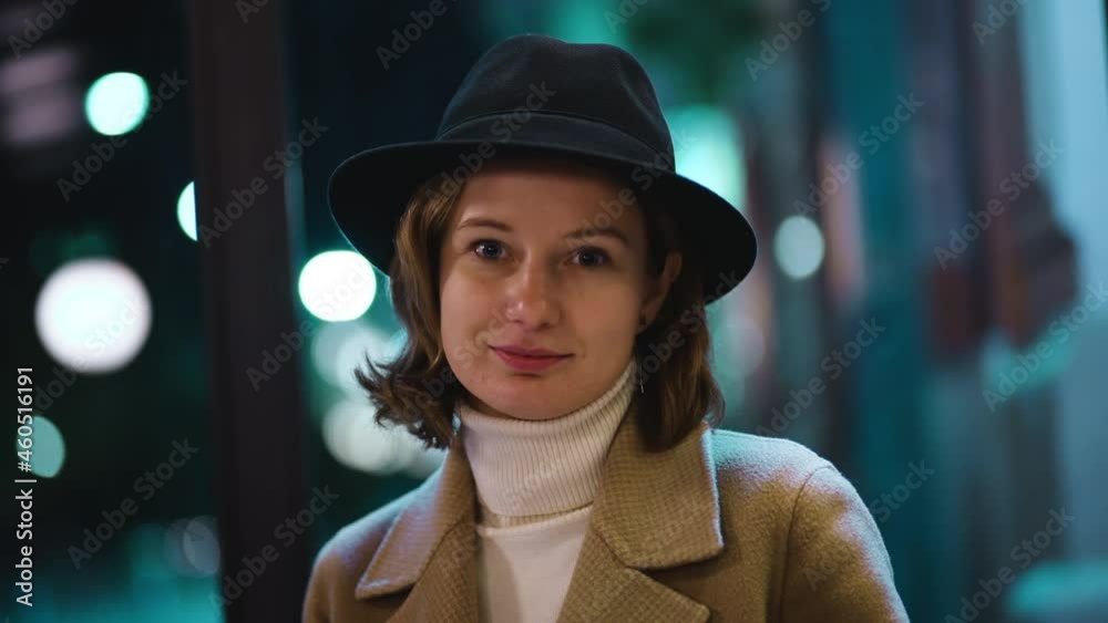 Cute girl in coat warm sweater and hat looks at camera and smiles and looks away. Tender and fragile girl on background of blurred night city with lights.Modesty and flirtation in eyes of stylish lady Cute girl in coat warm sweater and hat looks at camera and smiles and looks away. Tender and fragile girl on background of blurred night city with lights.Modesty and flirtation in eyes of stylish lady