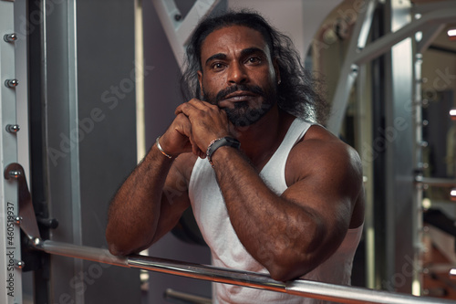 a black muscular man in a narrow white T-shirt stands at the gym and looks away. High quality photo
