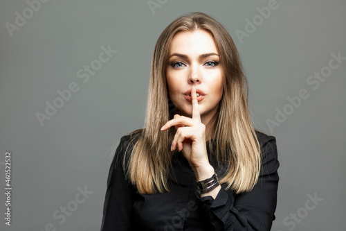 Young woman shows a sign of silence. A striking blonde in a black shirt put her finger to her lips. Silence and confidentiality. Gray background. photo