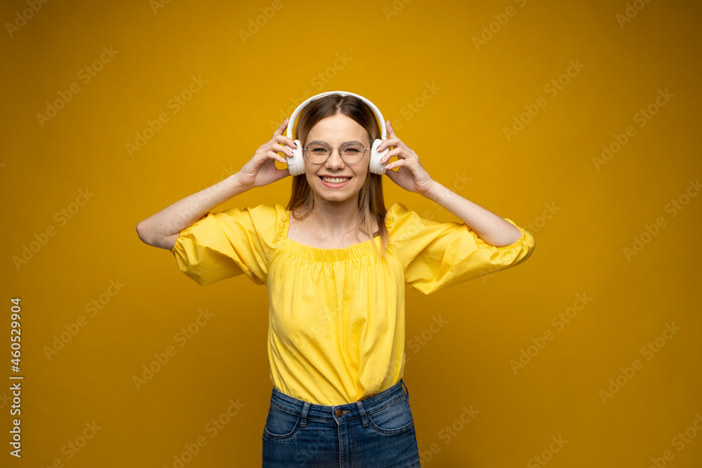 Beautiful attractive young blond woman wearing yellow t-shirt and glasses in white headphones listening music, dancing and laughing on blue background in studio. Relaxing and enjoying. Lifestyle. Beautiful attractive young blond woman wearing yellow t-shirt and glasses in white headphones listening music, dancing and laughing on blue background in studio. Relaxing and enjoying. Lifestyle.