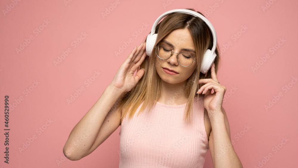 Beautiful attractive young blond woman wearing pink t-shirt and glasses in white headphones listening music and smiling on pink background in studio. Relaxing and enjoying. Lifestyle. Beautiful attractive young blond woman wearing pink t-shirt and glasses in white headphones listening music and smiling on pink background in studio. Relaxing and enjoying. Lifestyle.