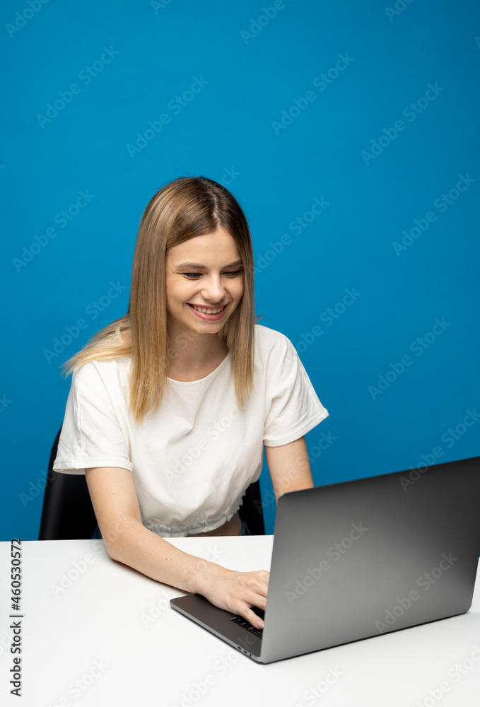 Portrait of a pretty young woman studying while sitting at the table with grey laptop computer, notebook. Smiling business woman working with a laptop isolated on a blue background. Portrait of a pretty young woman studying while sitting at the table with grey laptop computer, notebook. Smiling business woman working with a laptop isolated on a blue background.