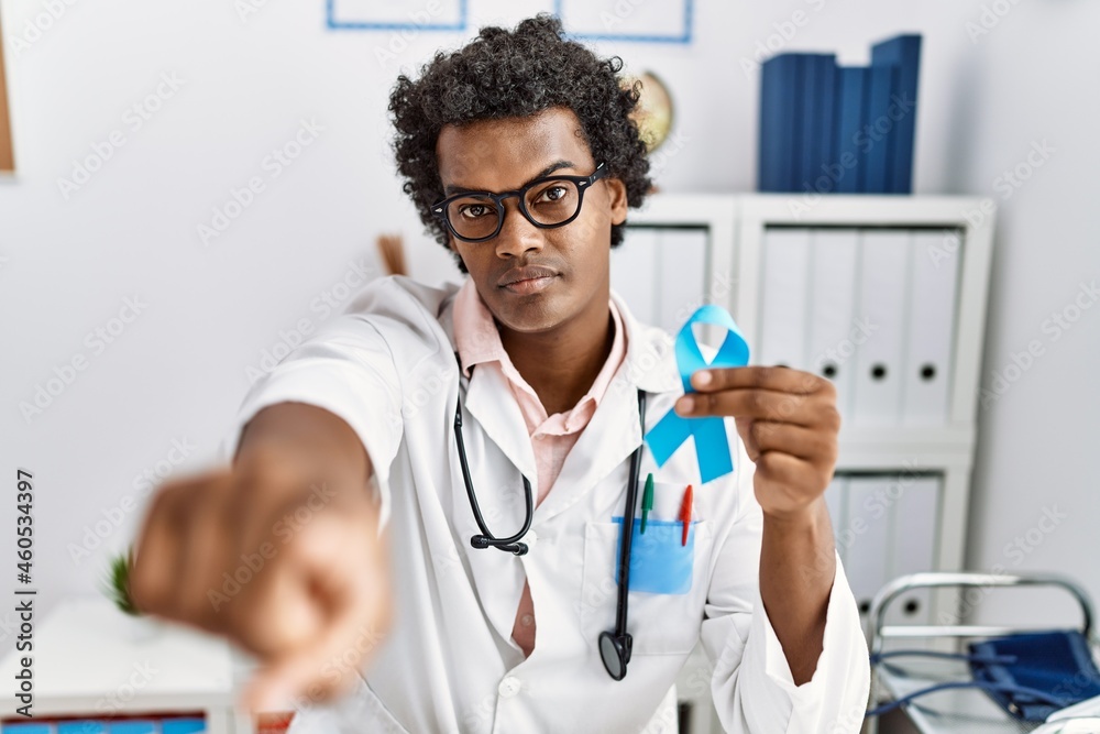 African doctor man holding blue ribbon pointing with finger to the camera and to you, confident gesture looking serious African doctor man holding blue ribbon pointing with finger to the camera and to you, confident gesture looking serious