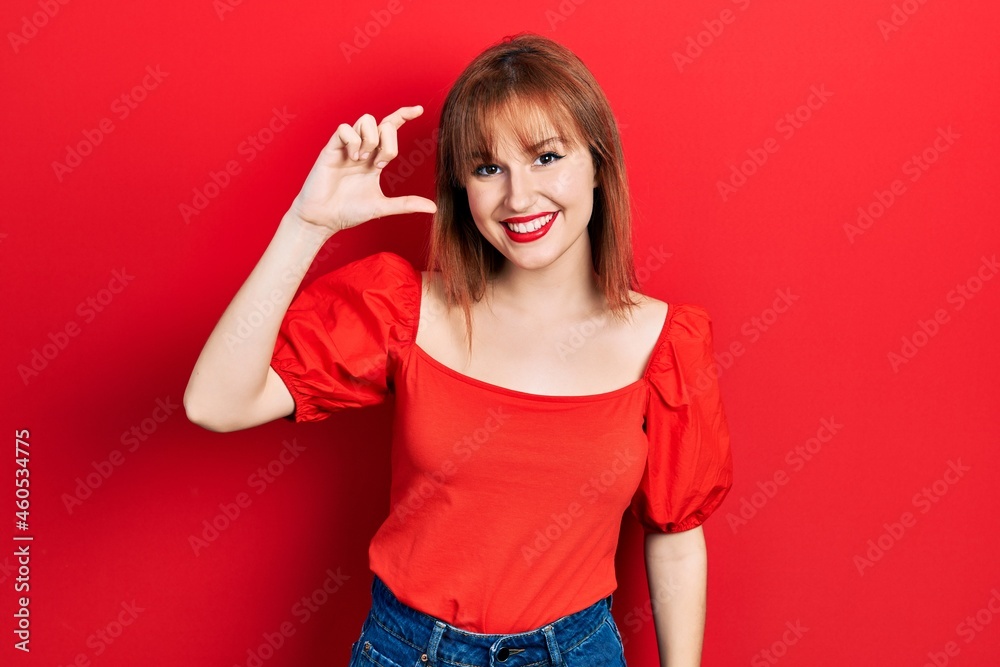 Redhead young woman wearing casual red t shirt smiling and confident gesturing with hand doing small size sign with fingers looking and the camera. measure concept. Redhead young woman wearing casual red t shirt smiling and confident gesturing with hand doing small size sign with fingers looking and the camera. measure concept.