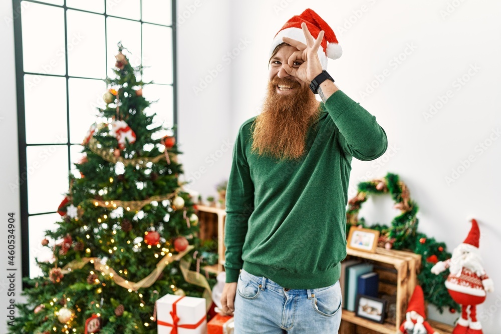 Redhead man with long beard wearing christmas hat by christmas tree doing ok gesture with hand smiling, eye looking through fingers with happy face. Redhead man with long beard wearing christmas hat by christmas tree doing ok gesture with hand smiling, eye looking through fingers with happy face.