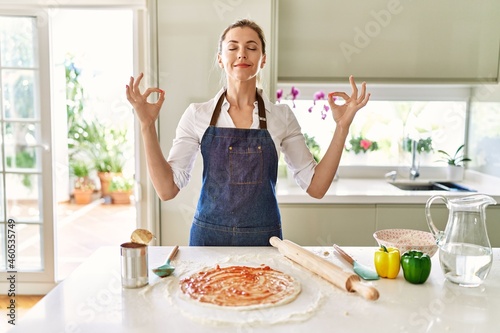 Beautiful blonde woman wearing apron cooking pizza relax and smiling with eyes closed doing meditation gesture with fingers. yoga concept.