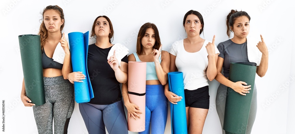 Obraz premium Group of women holding yoga mat standing over isolated background pointing up looking sad and upset, indicating direction with fingers, unhappy and depressed.