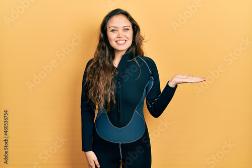 Schilderij op canvas Young hispanic girl wearing diver neoprene uniform smiling cheerful presenting and pointing with palm of hand looking at the camera