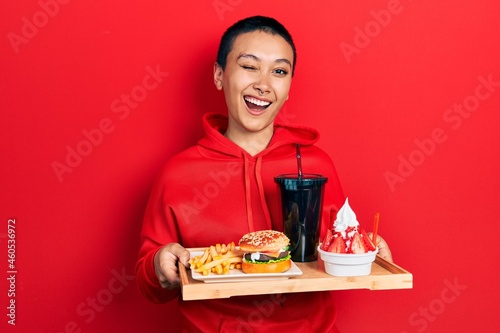 Beautiful hispanic woman with short hair eating a tasty classic burger with fries and soda winking looking at the camera with sexy expression  cheerful and happy face.