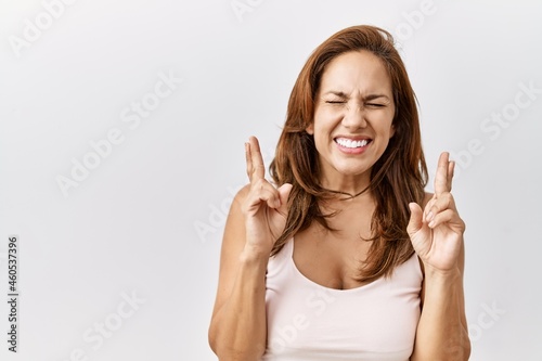 Middle age hispanic woman standing over isolated background gesturing finger crossed smiling with hope and eyes closed. luck and superstitious concept.