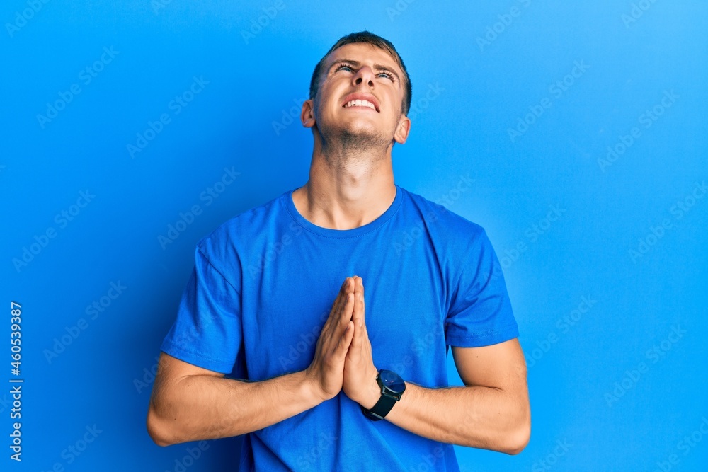 Young caucasian man wearing casual blue t shirt begging and praying with hands together with hope expression on face very emotional and worried. begging. Young caucasian man wearing casual blue t shirt begging and praying with hands together with hope expression on face very emotional and worried. begging.