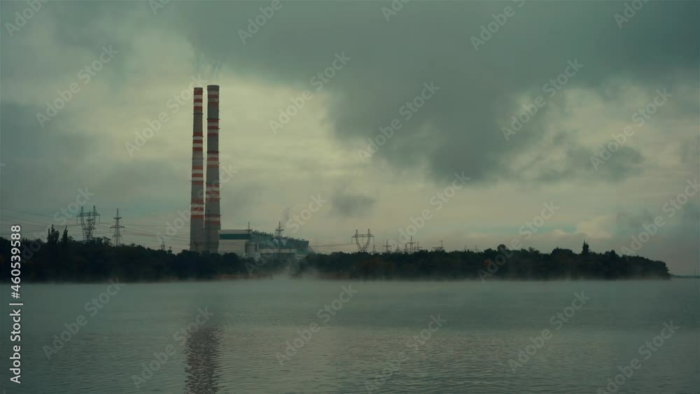 Power station on the lake shore in the evening. Steam or haze is spreading across the lake. Smoke is coming from the pipes, creating fog around and dark clouds. Around the forest because of which th Power station on the lake shore in the evening. Steam or haze is spreading across the lake. Smoke is coming from the pipes, creating fog around and dark clouds. Around the forest because of which th