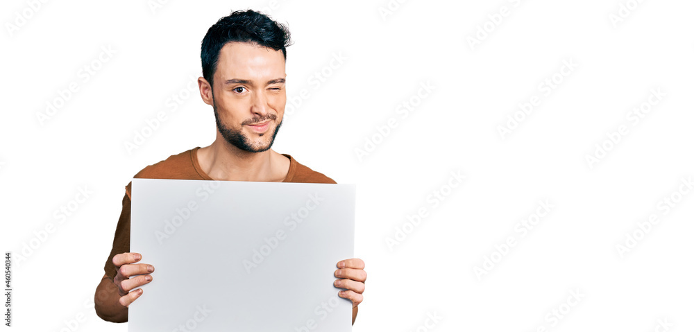 Hispanic man with beard holding blank empty banner winking looking at the camera with sexy expression, cheerful and happy face. Hispanic man with beard holding blank empty banner winking looking at the camera with sexy expression, cheerful and happy face.