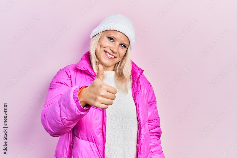 Beautiful caucasian blonde woman wearing wool hat and winter coat doing happy thumbs up gesture with hand. approving expression looking at the camera showing success. Beautiful caucasian blonde woman wearing wool hat and winter coat doing happy thumbs up gesture with hand. approving expression looking at the camera showing success.