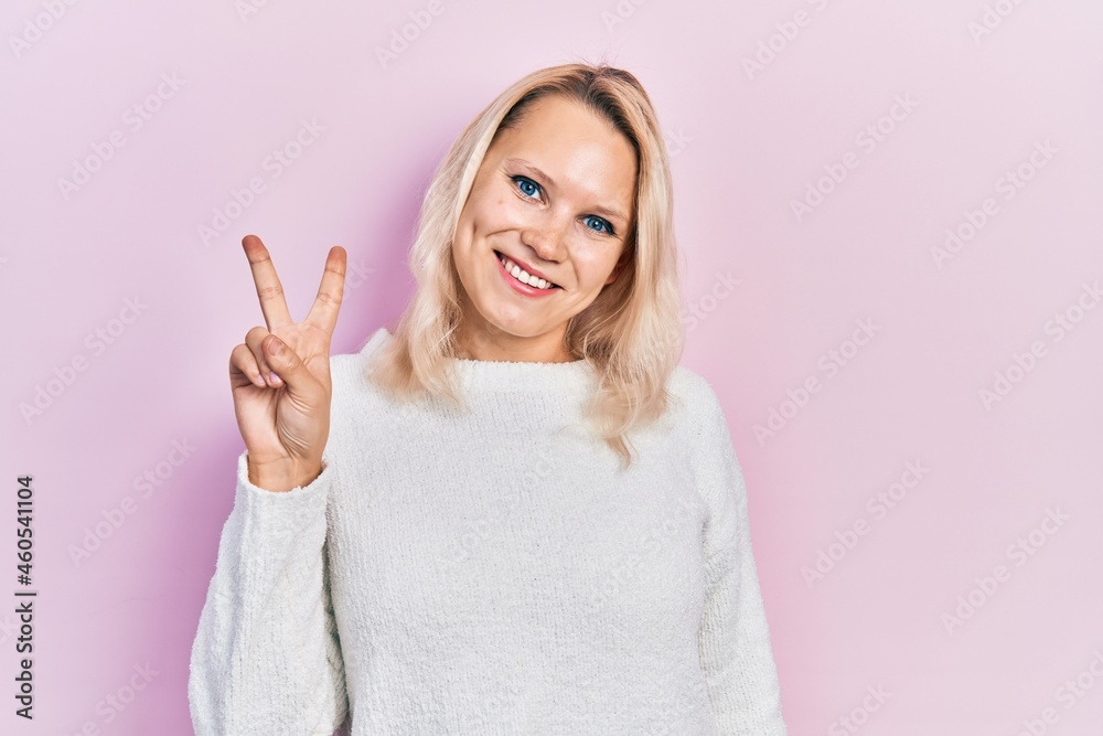 Beautiful caucasian blonde woman wearing casual winter sweater smiling with happy face winking at the camera doing victory sign. number two. Beautiful caucasian blonde woman wearing casual winter sweater smiling with happy face winking at the camera doing victory sign. number two.