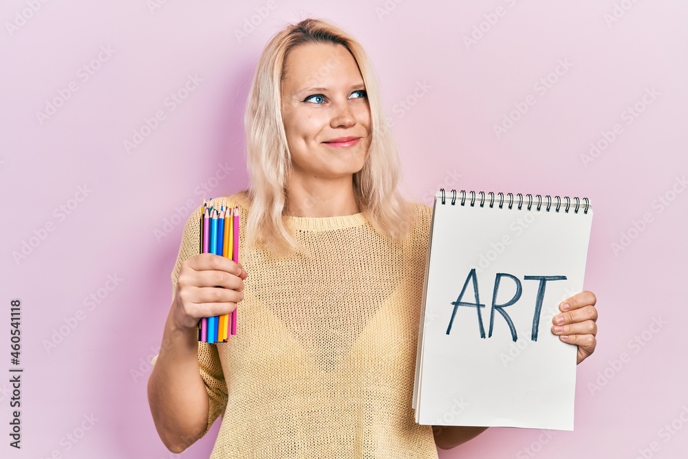 Beautiful caucasian blonde woman holding art notebook and color pencils smiling looking to the side and staring away thinking. Beautiful caucasian blonde woman holding art notebook and color pencils smiling looking to the side and staring away thinking.