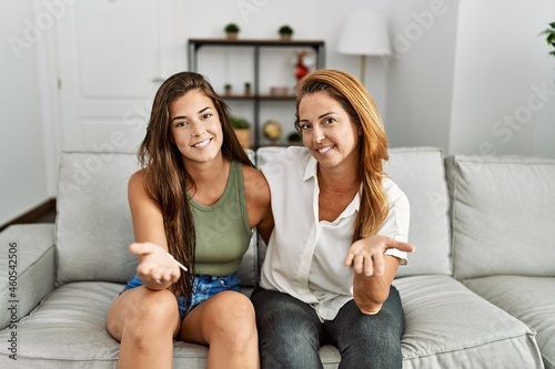 Mother and daughter together sitting on the sofa at home smiling cheerful offering palm hand giving assistance and acceptance.