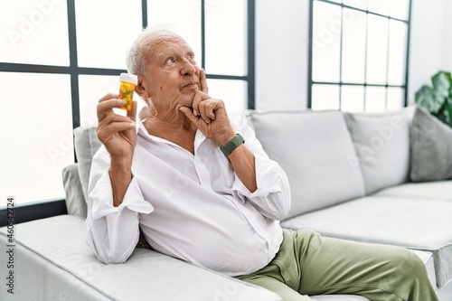 Senior man holding pills with hand on chin thinking about question  pensive expression. smiling and thoughtful face. doubt concept.