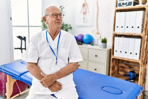 Senior physiotherapy man working at pain recovery clinic smiling looking to the side and staring away thinking.