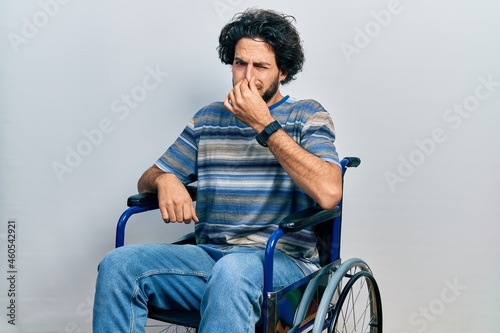 Handsome hispanic man sitting on wheelchair smelling something stinky and disgusting, intolerable smell, holding breath with fingers on nose. bad smell