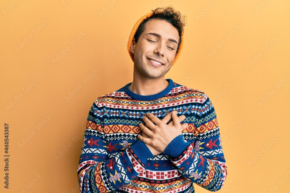 Fototapeta premium Young handsome man wearing wool hat and colorful sweater smiling with hands on chest with closed eyes and grateful gesture on face. health concept.