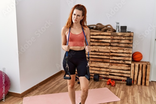 Young redhead woman training with elastic arm bands clueless and confused expression. doubt concept.