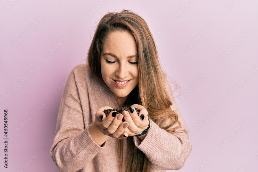 Young woman holding raw coffee beans and smelling roasted aroma over pink background
