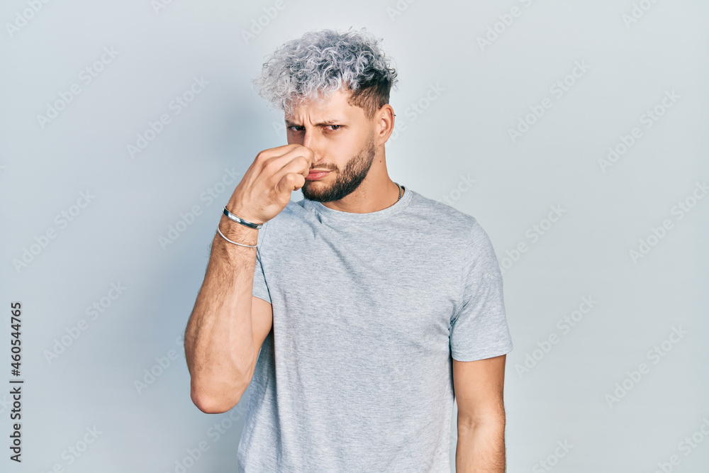 Young hispanic man with modern dyed hair wearing casual grey t shirt smelling something stinky and disgusting, intolerable smell, holding breath with fingers on nose. bad smell Young hispanic man with modern dyed hair wearing casual grey t shirt smelling something stinky and disgusting, intolerable smell, holding breath with fingers on nose. bad smell
