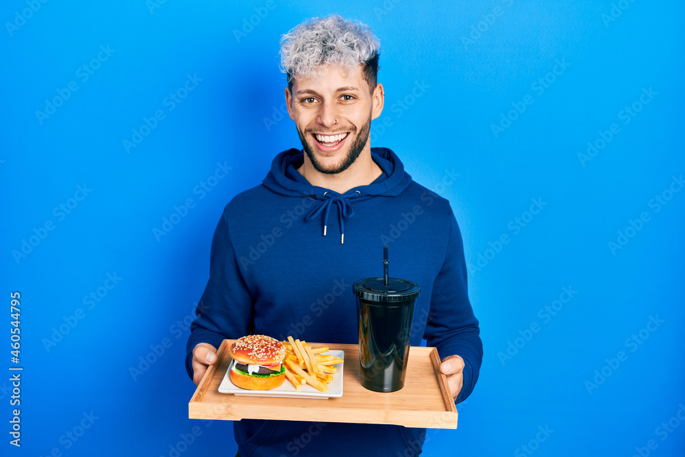 Young hispanic man with modern dyed hair eating a tasty classic burger with fries and soda smiling and laughing hard out loud because funny crazy joke. Young hispanic man with modern dyed hair eating a tasty classic burger with fries and soda smiling and laughing hard out loud because funny crazy joke.