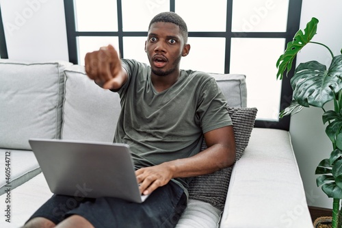 Young african american man using laptop at home sitting on the sofa pointing with finger surprised ahead, open mouth amazed expression, something on the front