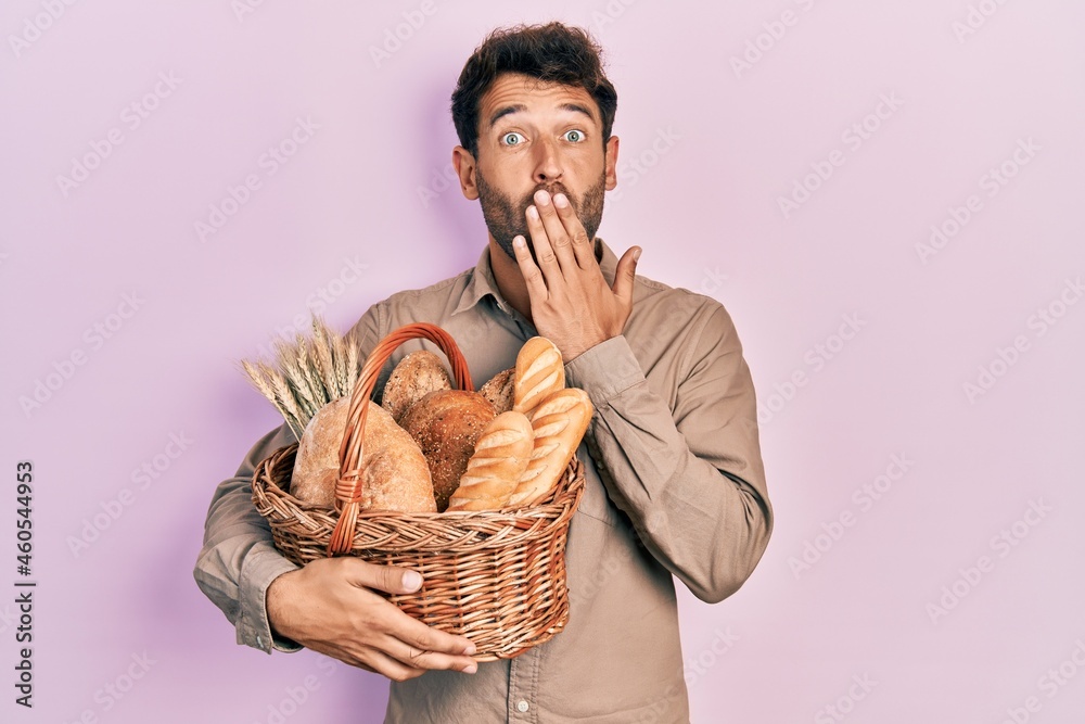 Handsome man with beard holding wicker basket with bread covering mouth with hand, shocked and afraid for mistake. surprised expression Handsome man with beard holding wicker basket with bread covering mouth with hand, shocked and afraid for mistake. surprised expression