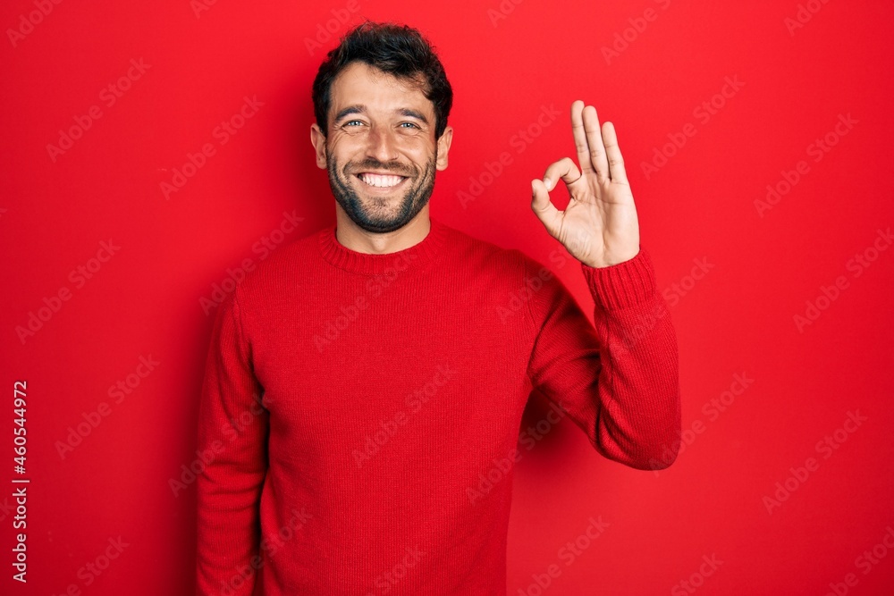 Handsome man with beard wearing casual red sweater smiling positive doing ok sign with hand and fingers. successful expression. Handsome man with beard wearing casual red sweater smiling positive doing ok sign with hand and fingers. successful expression.
