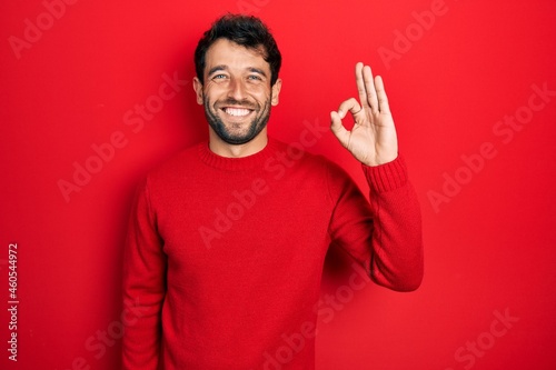 Handsome man with beard wearing casual red sweater smiling positive doing ok sign with hand and fingers. successful expression.