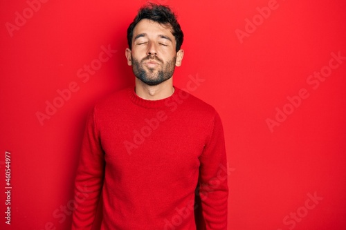 Handsome man with beard wearing casual red sweater looking at the camera blowing a kiss on air being lovely and sexy. love expression.