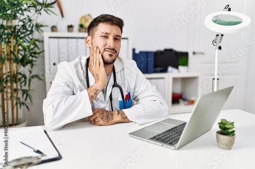 Young doctor working at the clinic using computer laptop touching mouth with hand with painful expression because of toothache or dental illness on teeth. dentist