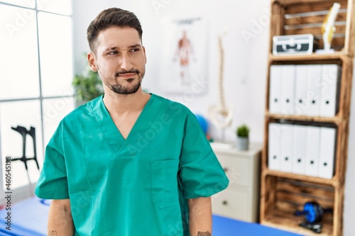 Young physiotherapist man working at pain recovery clinic smiling looking to the side and staring away thinking.