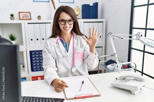 Young doctor woman wearing doctor uniform and stethoscope at the clinic showing and pointing up with fingers number five while smiling confident and happy.