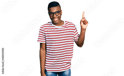 Young african american man wearing casual t shirt and glasses showing and pointing up with finger number one while smiling confident and happy.