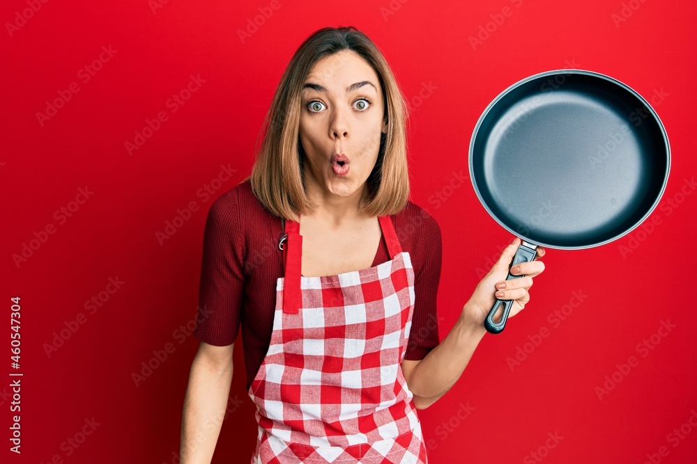 Young caucasian blonde woman wearing cook apron holding pan scared and amazed with open mouth for surprise, disbelief face Young caucasian blonde woman wearing cook apron holding pan scared and amazed with open mouth for surprise, disbelief face