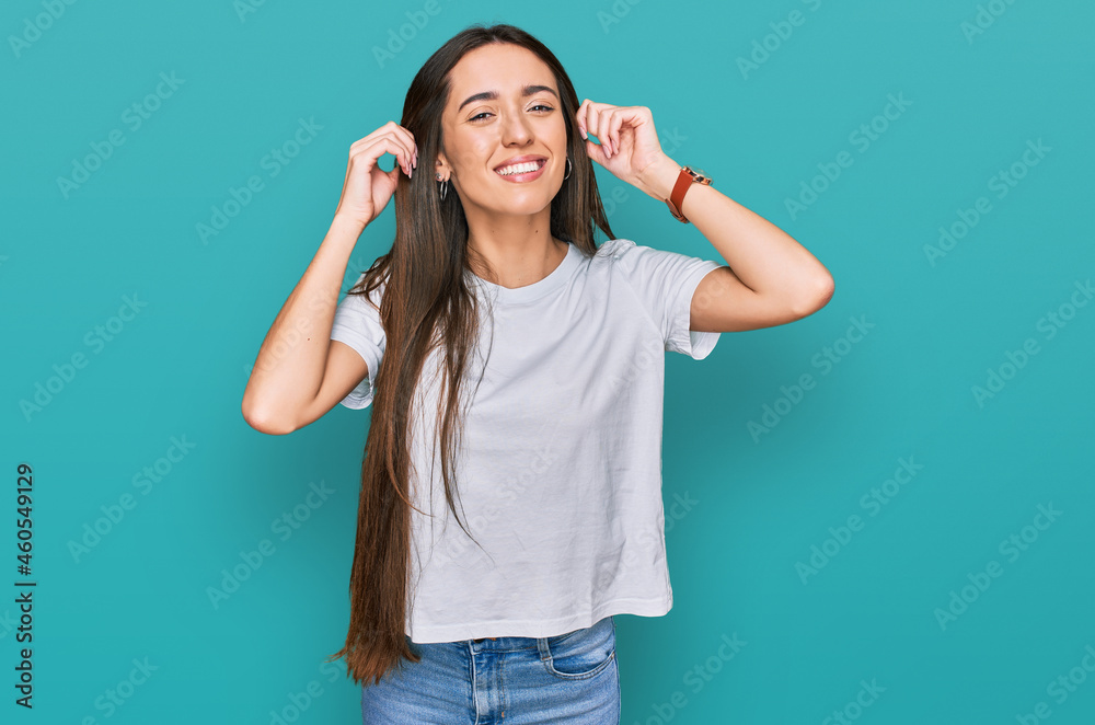 Fototapeta premium Young hispanic girl wearing casual white t shirt smiling pulling ears with fingers, funny gesture. audition problem