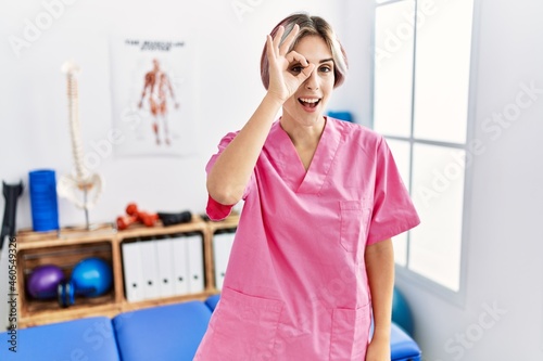 Young nurse woman working at pain recovery clinic smiling happy doing ok sign with hand on eye looking through fingers