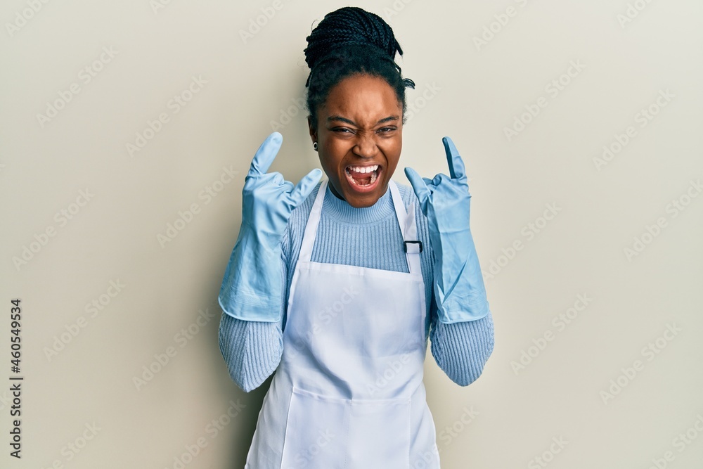 African american woman with braided hair wearing cleaner apron and gloves shouting with crazy expression doing rock symbol with hands up. music star. heavy concept. African american woman with braided hair wearing cleaner apron and gloves shouting with crazy expression doing rock symbol with hands up. music star. heavy concept.