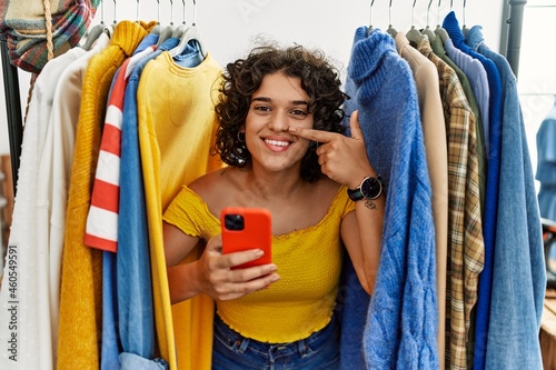 Young hispanic woman searching clothes on clothing rack using smartphone pointing with hand finger to face and nose, smiling cheerful. beauty concept