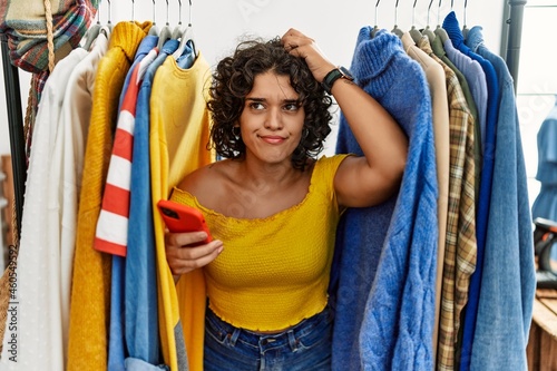 Young hispanic woman searching clothes on clothing rack using smartphone confuse and wondering about question. uncertain with doubt, thinking with hand on head. pensive concept.