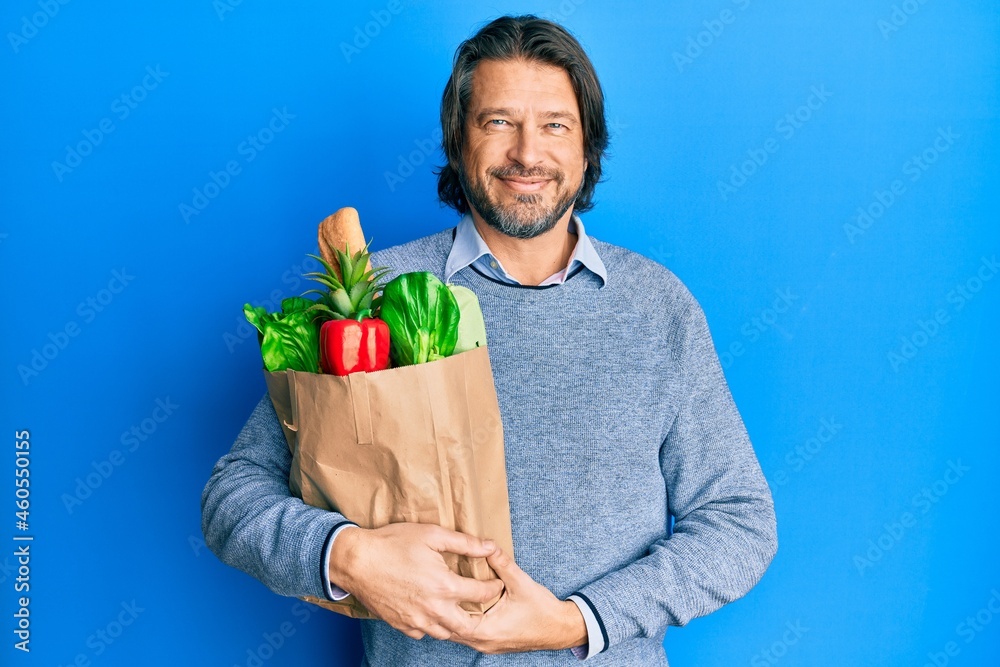Middle age handsome man holding paper bag with groceries looking positive and happy standing and smiling with a confident smile showing teeth