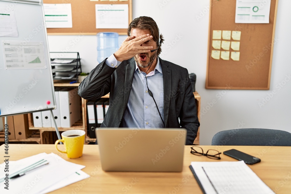 Handsome middle age man wearing call center agent headset at the office peeking in shock covering face and eyes with hand, looking through fingers with embarrassed expression.